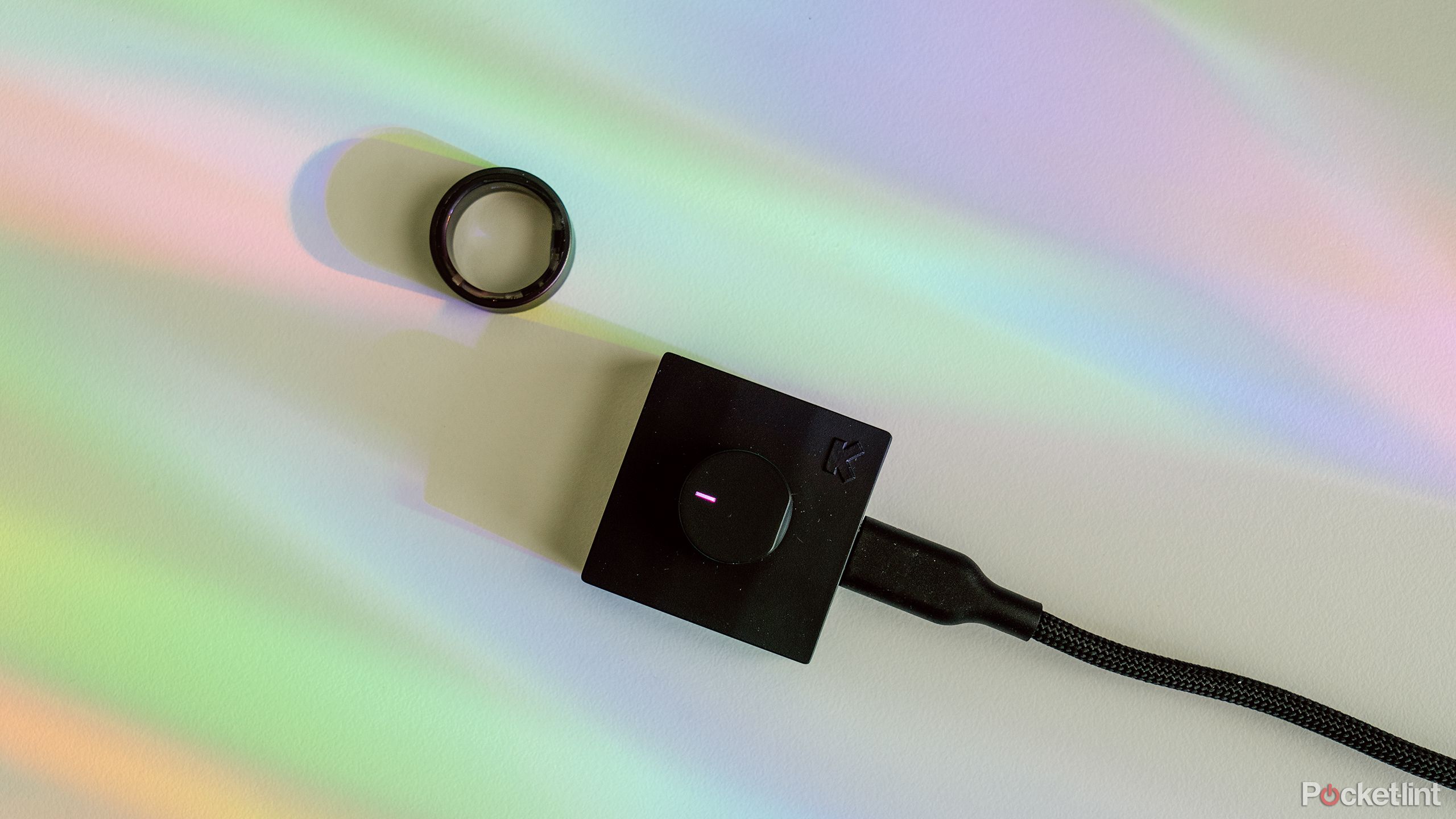 Ultrahuman Ring Air در کنار شارژر خود روی یک میز سفید با نور رنگین کمان. 