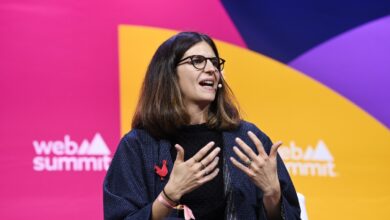 A brown haired woman wearing glasses stands in front of the a multi-colored background with the web summit logo