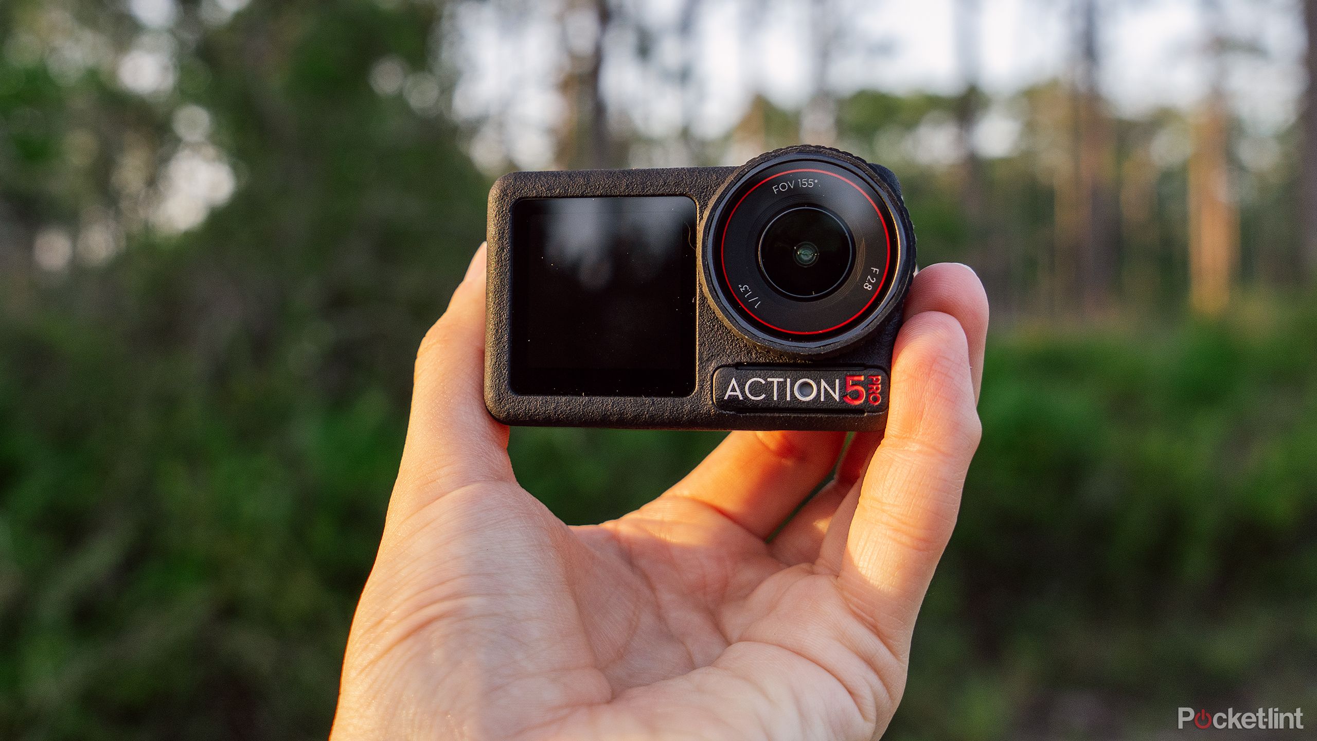 یک دست DJI Osmo Action 5 Pro را در مقابل جنگلی تار نگه می دارد. 
