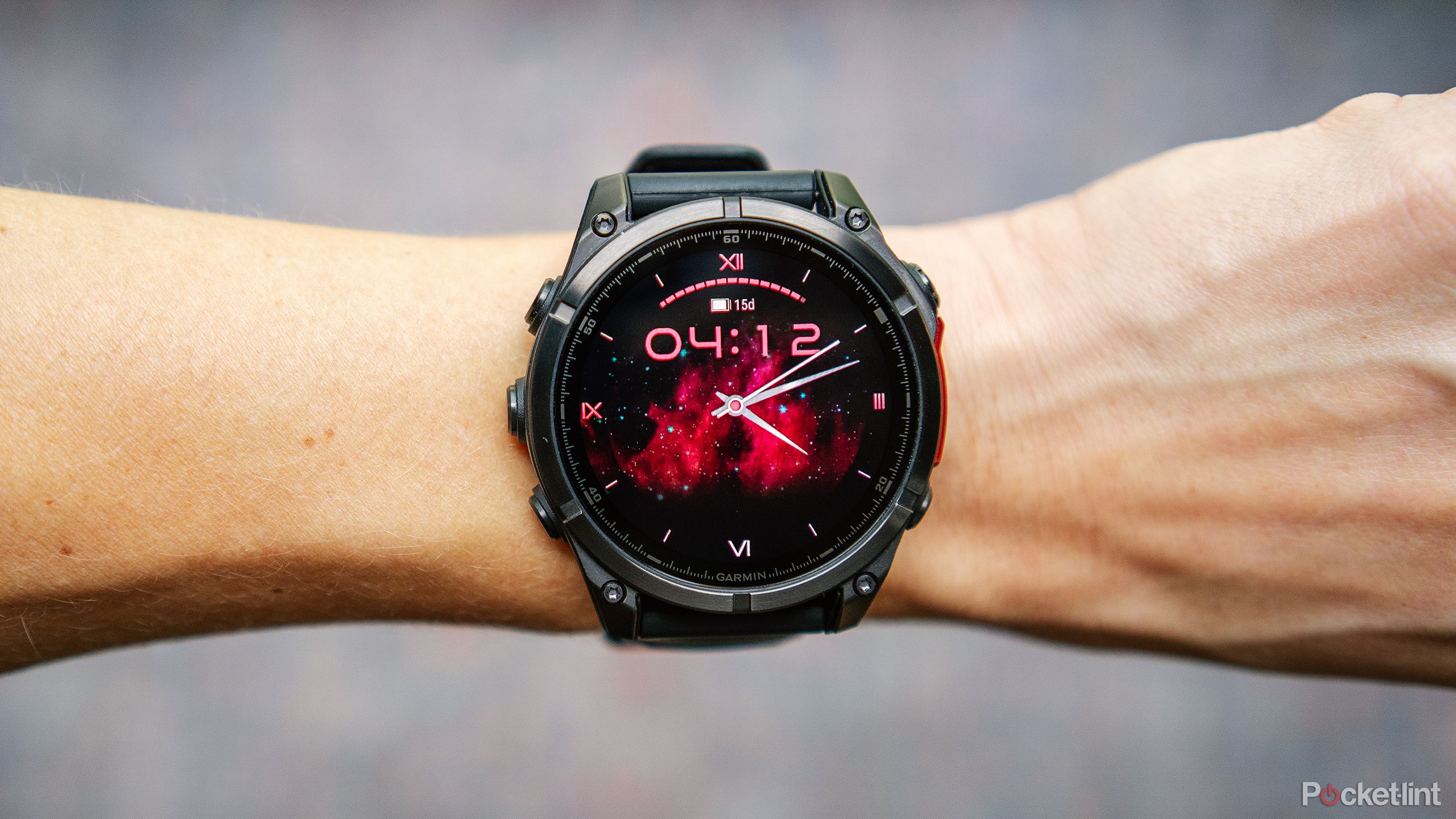 Garmin Fenix ​​8 AMOLED با صفحه ساعت Ad Astra روی مچ دستی بالای پس‌زمینه آبی و قرمز تار قرار دارد. 