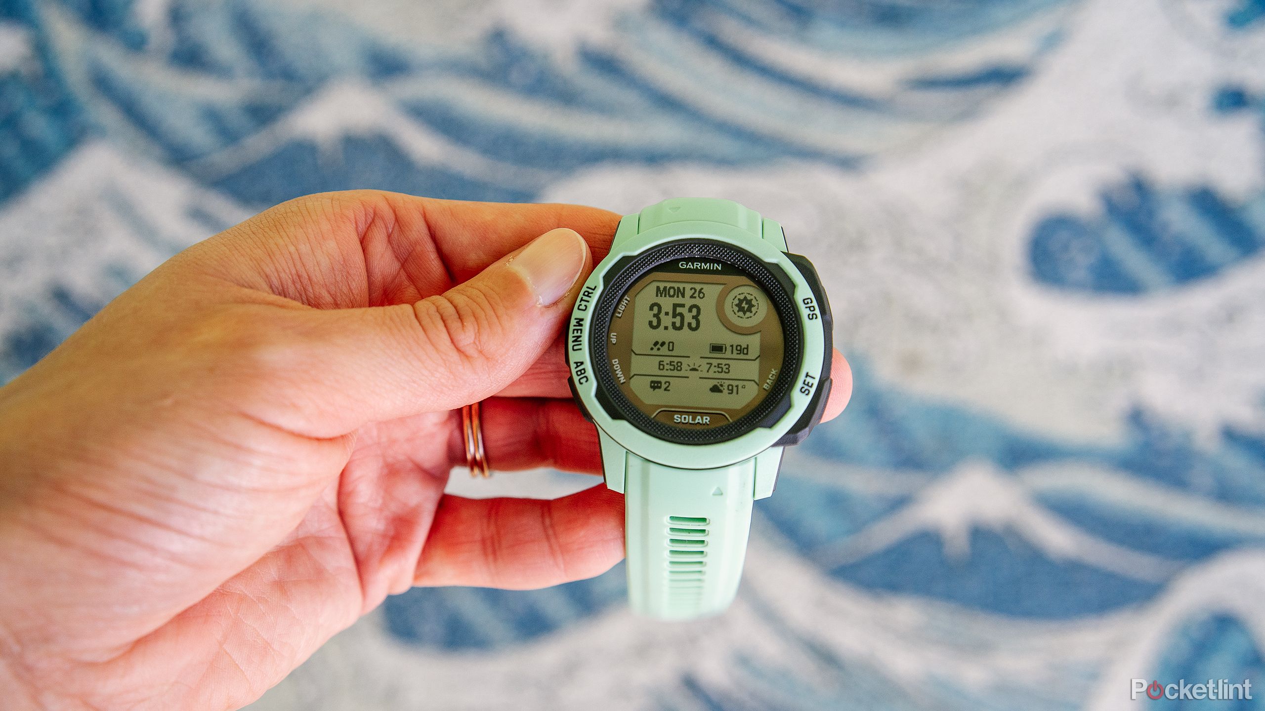 یک دست نعناع Garmin Instinct 2S را بالای یک تصویر تار از امواج نگه می دارد. 