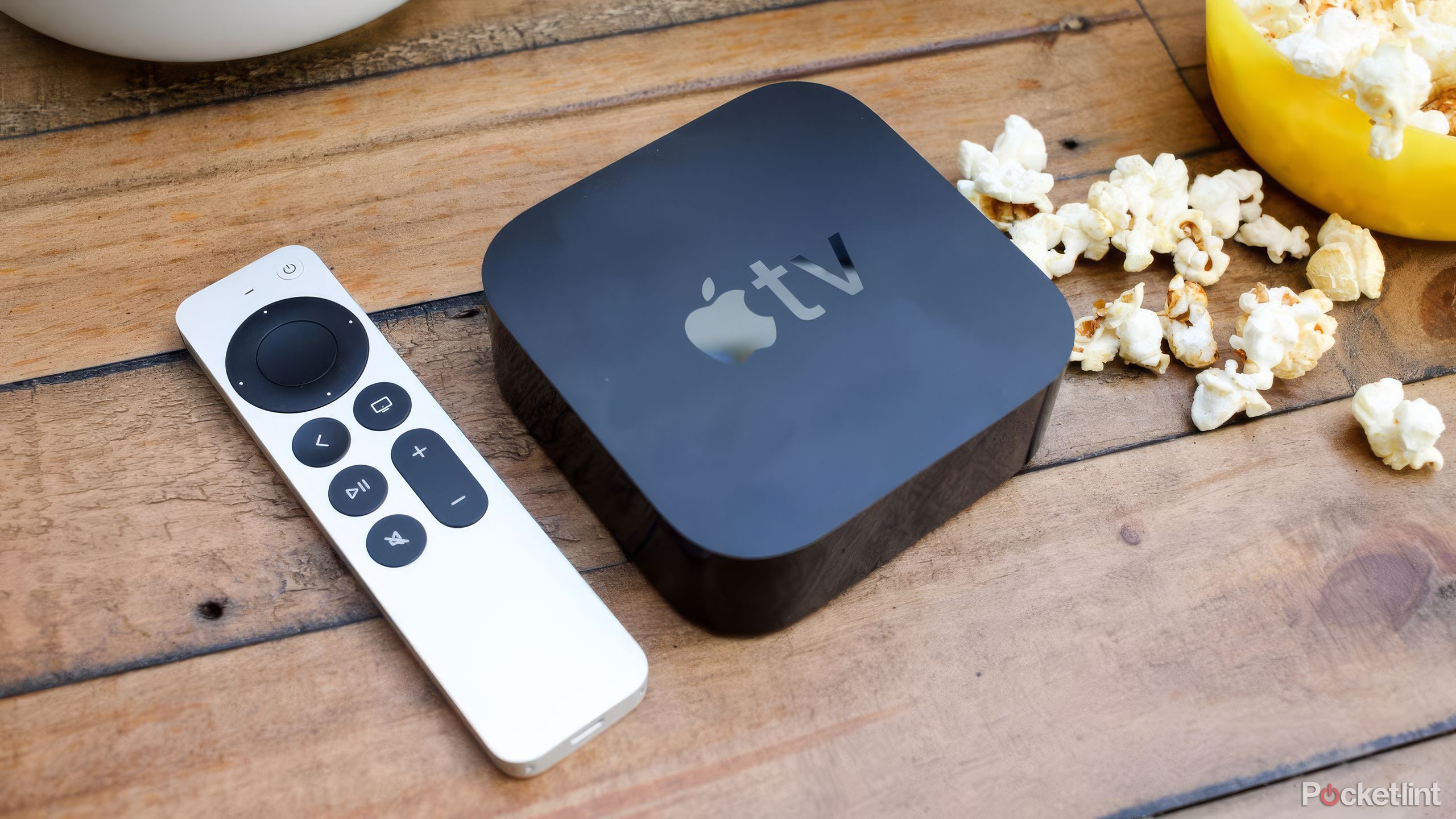یک کنترل و جعبه Apple TV روی یک میز چوبی در کنار پاپ کورن قرار دارد.