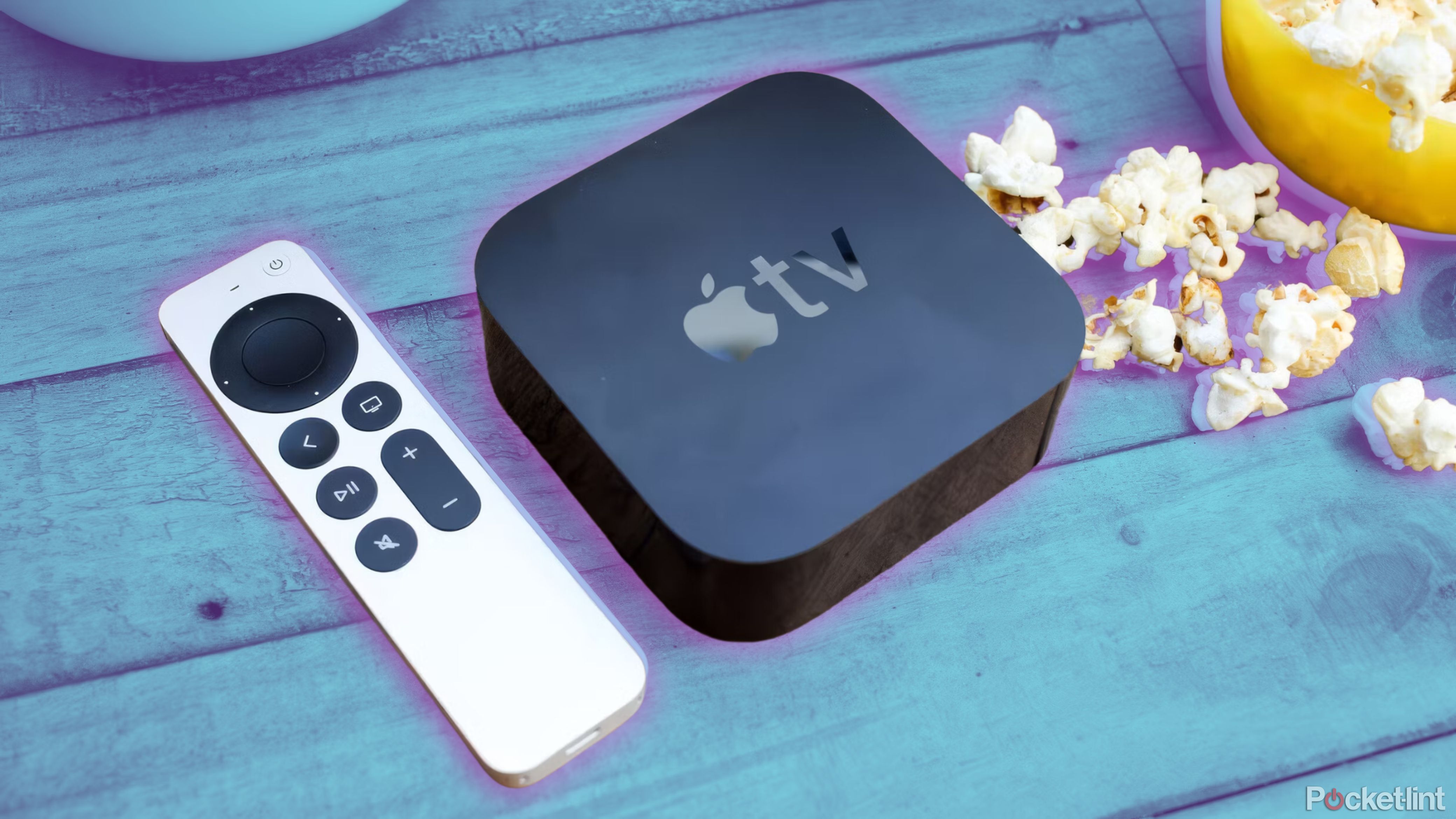 یک کنترل و جعبه Apple TV روی یک میز چوبی در کنار پاپ کورن قرار دارد.
