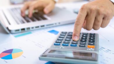 person with one hand typing on a laptop and the other working a calculator