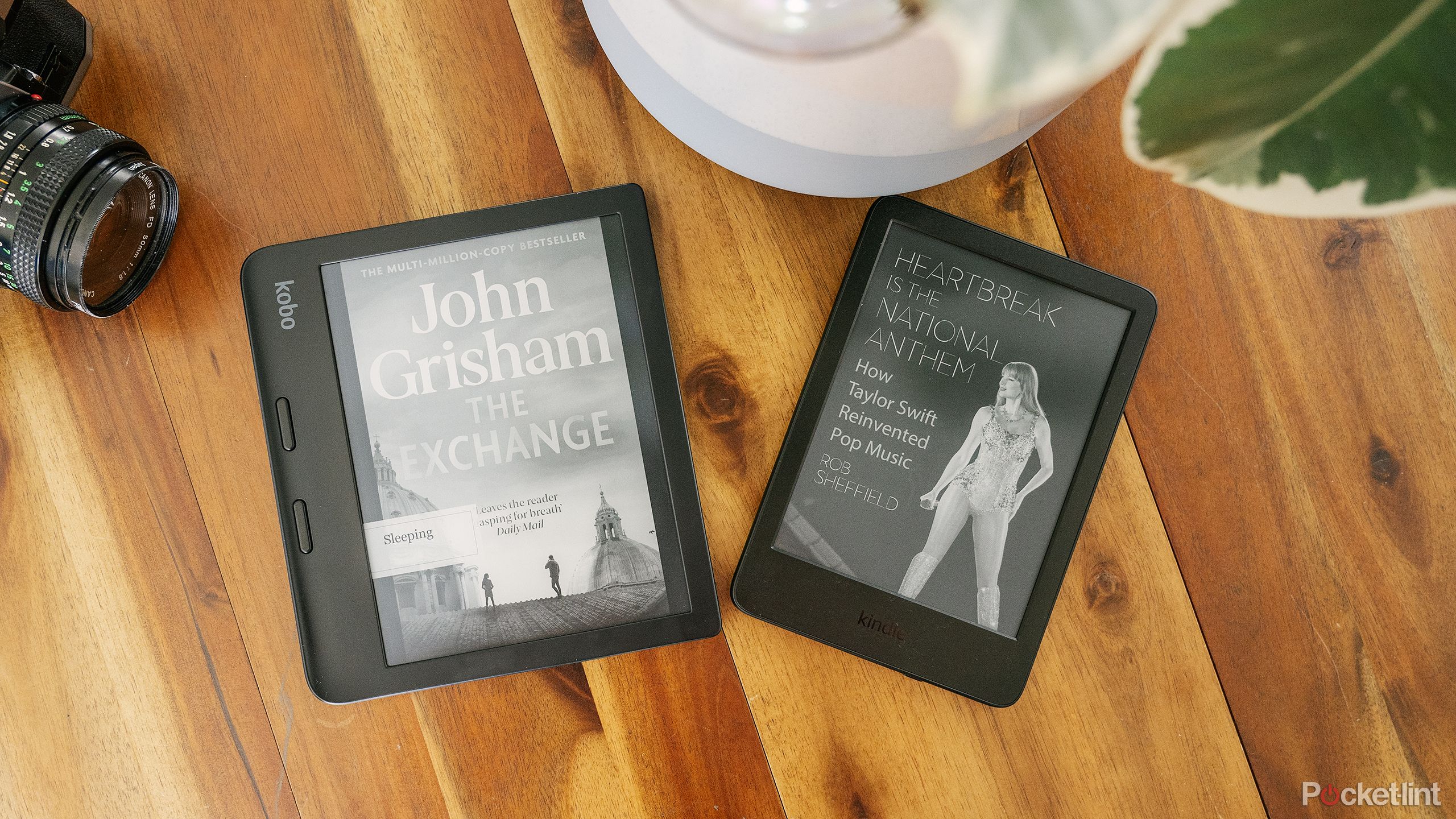Kindle Paperwhite در کنار Kobo Libra 2 روی میز چوبی در کنار یک گیاه گلدانی و دوربین قدیمی قرار دارد.