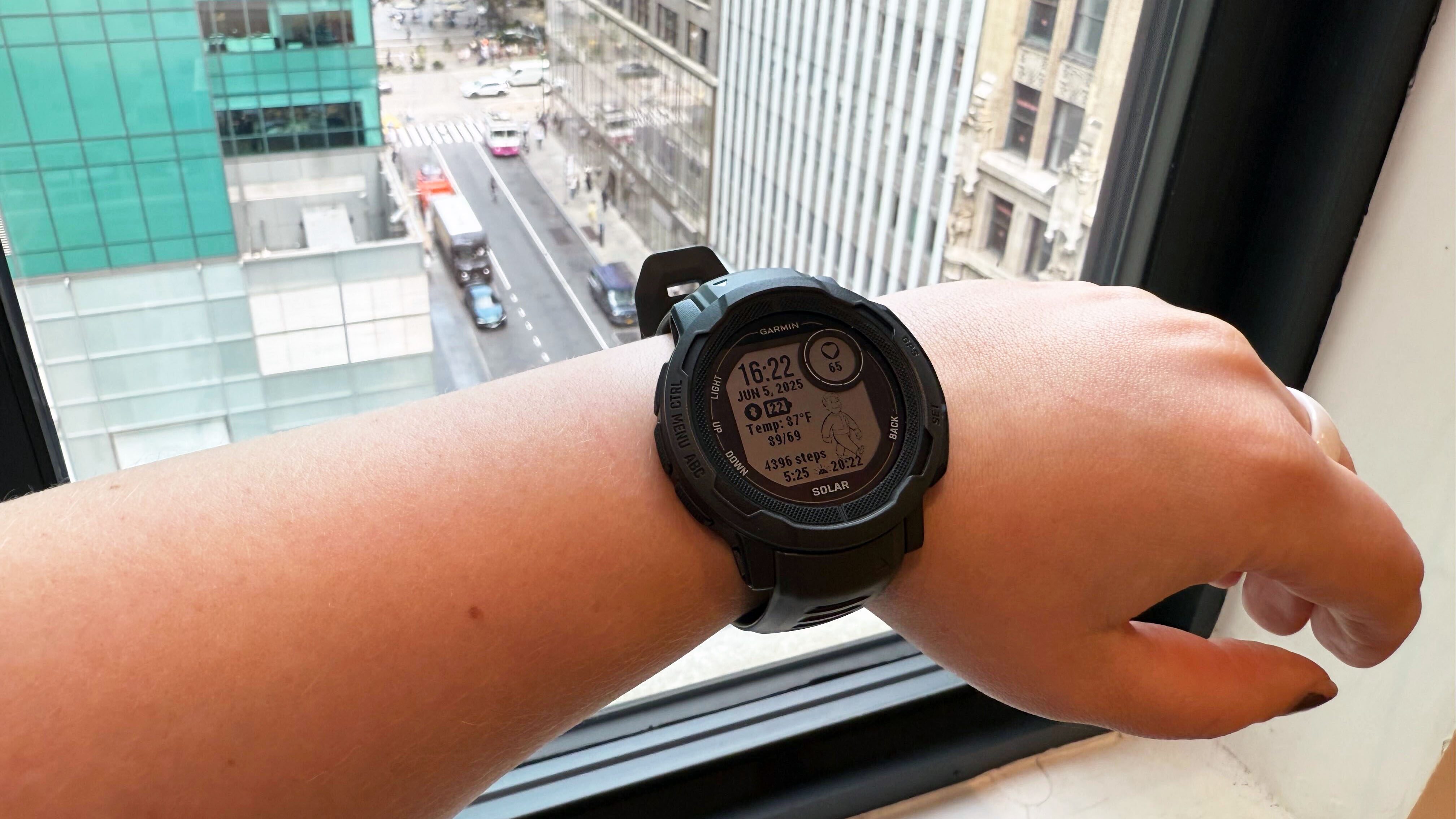 Garmin Instinct 2 Solar در مچ دست چپ ، جلوی پنجره ای مشرف به پارک برایانت در شهر نیویورک.