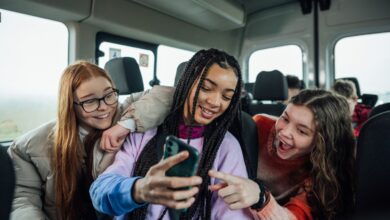 A front view of a group of three teenage girls on the minibus on the way to go on a hiking field trip. They are looking at one of the girls mobile phones and smiling and pointing at the screen.